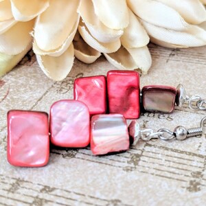 May include: A pair of earrings with pink and red square-shaped beads and silver hooks. The beads have a pearlescent finish.