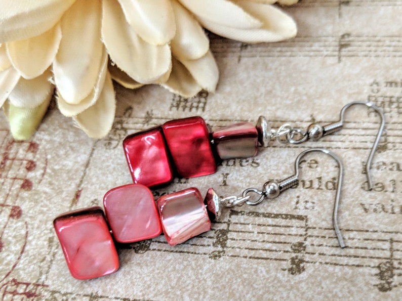 May include: A pair of red and pink shell earrings with silver hooks. The earrings are made of stacked square-shaped shells.
