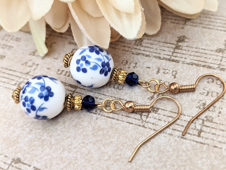 May include: A pair of blue and white porcelain earrings with gold accents and blue crystal beads. The earrings are on a gold background.