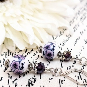 May include: Dangle earrings with white ceramic beads decorated with a purple floral design. The earrings have silver-coloured metal accents and dark purple crystal beads. The earrings are set against a white background with a flower.