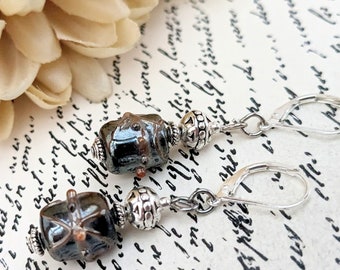 May include: A pair of drop earrings featuring dark, metallic-toned square beads with copper-coloured accents. Each earring includes a silver-toned decorative spacer bead and a silver-toned leverback closure, creating a balanced and detailed design.