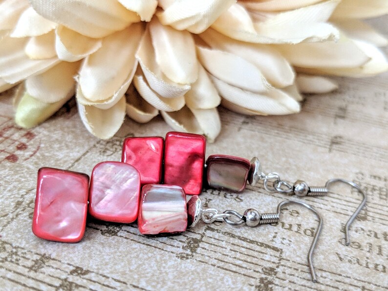 May include: Pink and red square shaped earrings with silver hooks. The earrings are made of shell and have a pearlescent finish.