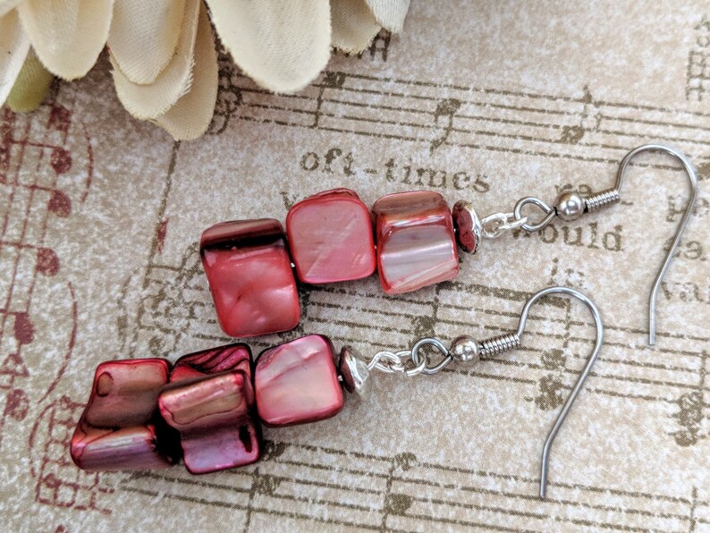 May include: A pair of red and silver earrings with a teardrop shape. The earrings are made of red shell beads and silver hooks.