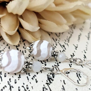 May include: Close-up of dangle earrings featuring large, white and gray striped beads. The earrings have silver-colored metal accents and lever-back closures. The background includes a soft, cream-colored flower and script-style writing.