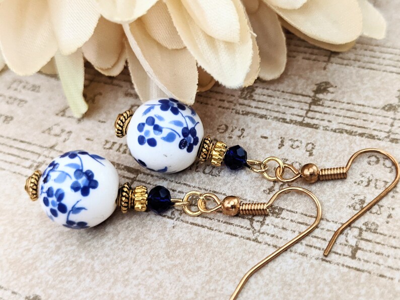 May include: A pair of dangle earrings with blue and white porcelain beads and gold accents. The beads feature a floral design.