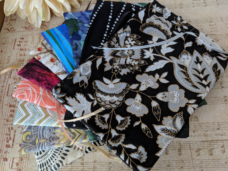 May include: A stack of fabric pouches with various patterns, including floral, geometric, and abstract designs. The pouches are made of different fabrics, including cotton and linen. The pouches are tied together with a ribbon.
