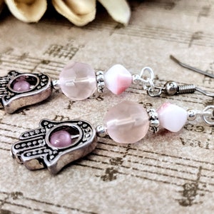 May include: Pink and silver earrings featuring a hamsa hand charm, rose quartz beads, and white shell beads.