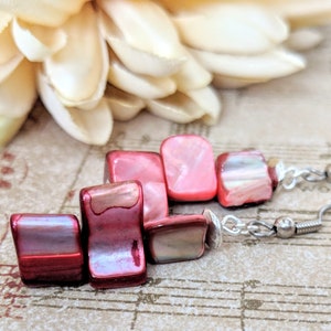 May include: A pair of dangle earrings with red and pink iridescent shell beads. The earrings have silver hooks.