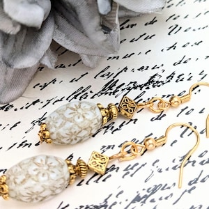 May include: Close-up of a pair of dangle earrings. Each earring features a white, patterned, oval bead with gold-colored accents. The earrings have gold-colored hooks and decorative square and round elements. The background is a vintage-style paper.