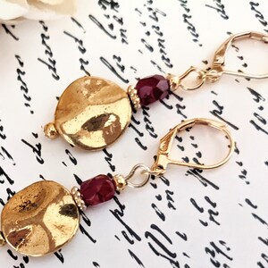 May include: Gold earrings with a hammered disc charm and a single red gemstone bead. The earrings have a lever back closure.