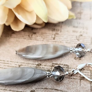 Puede incluir: Un par de pendientes de plata con un colgante de concha de forma ovalada gris y una cuenta de cristal transparente. Los pendientes tienen un cierre de palanca.