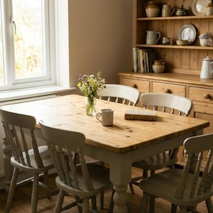 May include: A rectangular wooden dining table with a natural light brown top and light grey painted legs, surrounded by four matching light grey spindle-back chairs. A glass vase with wildflowers and a ceramic mug sit on the table surface.