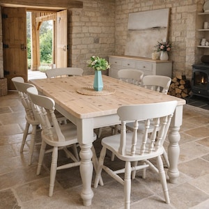 May include: A rectangular dining table with a natural light wood top and a cream-coloured base featuring turned legs. Six matching cream-coloured spindle-back chairs are arranged around the table. A light blue vase with green foliage sits on a round woven placemat in the centre of the table.