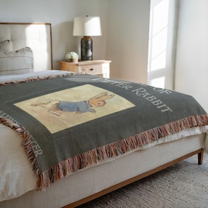 Puede incluir: Una manta de tejido verde con borde de flecos presenta una ilustración vintage de Peter Rabbit del libro "El cuento de Peter Rabbit".