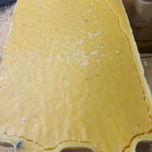 May include: Close-up of a large, rectangular block of yellow beeswax. The wax has a smooth, slightly textured surface with a few small imperfections. The edges of the block are uneven, and the overall appearance suggests a natural, unprocessed material. The color is a warm, golden yellow.