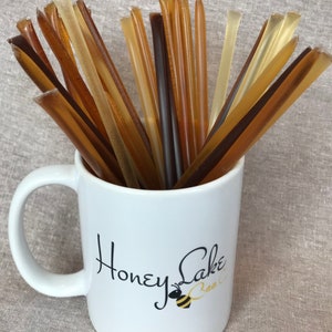 May include: A white ceramic mug with a black logo that says "Honey Lake Bee Co."  The mug is filled with a variety of honey sticks in different shades of brown and amber.