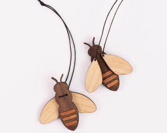 Pode incluir: Dois ornamentos de abelha de madeira com cordões pretos. As abelhas são marrons com asas de cor marrom claro e listras laranja.