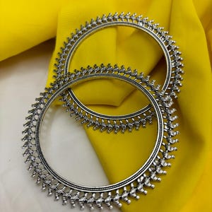 May include: Two silver bangles with intricate designs. The bangles are on a yellow fabric background.