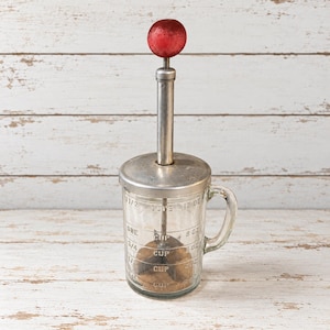 May include: A vintage glass kitchen beater with a clear measuring pitcher featuring markings for cups and ounces. It has a metal lid, a central vertical plunger mechanism, and a round red handle at the top. The base contains a metal whisk.
