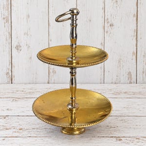 May include: A two-tiered serving stand featuring two circular gold-toned metal trays with beaded edges. The central support column is made of polished silver-toned metal with a ring handle at the top and a circular base at the bottom.