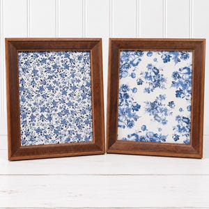 May include: Two rectangular picture frames with medium-brown wooden borders. Each frame contains a white background featuring a blue floral pattern. The left frame displays a dense, small-scale floral print, while the right frame shows a larger, more spaced-out blue rose design.
