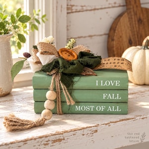 May include: A stack of three sage green books with white text reading I LOVE FALL MOST OF ALL. The books are topped with a burlap and dark green fabric bow, a wooden button, and a jute twine strand featuring three wooden beads and a tassel.