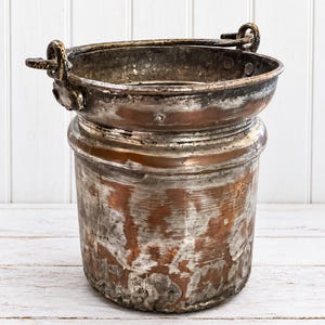 May include: A weathered copper bucket with a cylindrical body and a wide, flared rim. The exterior features a distressed, tarnished finish with visible patches of exposed copper and dark oxidation. It includes a sturdy metal swing handle.