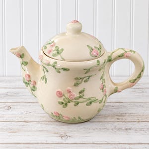 May include: A cream-colored ceramic teapot featuring a raised, three-dimensional vine pattern with small green leaves and delicate pink rosebuds. The teapot includes a matching lid with a knob handle, a curved spout, and a sturdy side handle.