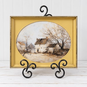 May include: A rectangular gold-toned frame with an oval inner border containing a landscape painting of a white cottage and a large tree. The artwork is displayed on a black metal stand with decorative scrollwork at the base and top.