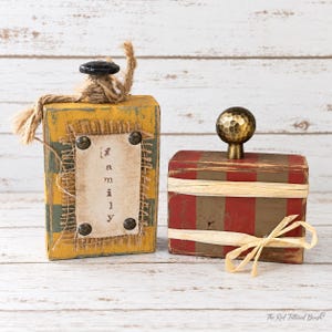 May include: Two rectangular wooden blocks. The left block is yellow with green stripes, topped with a black knob and jute twine, featuring a central tag with the word family. The right block is red with brown stripes, topped with a gold knob.