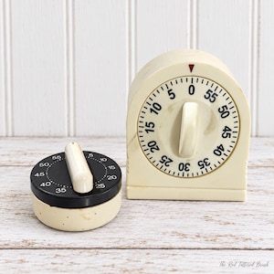 May include: Two kitchen timers: one round, low-profile timer with a black face and cream base, and one arched, upright timer with a cream face and black numerals. Both feature a central cream-colored knob and a red indicator triangle.