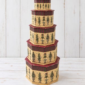 May include: A stack of five hexagonal nesting boxes in descending size order. Each box features a cream-colored base with a repeating pattern of green topiary trees in urns, topped with a dark red and green striped lid and matching base trim.