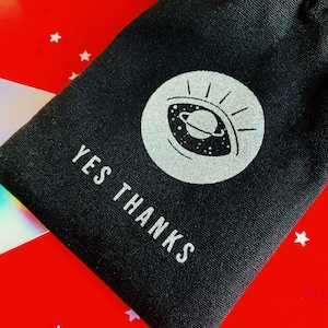 以下が含まれることがあります： 銀色の目のグラフィックと「YES THANKS」というテキストが銀色で印刷された黒いキャンバスポーチ