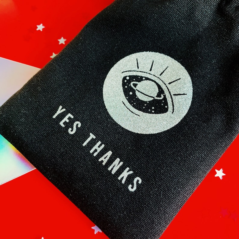 以下が含まれることがあります： 銀色の目のグラフィックと「YES THANKS」というテキストが銀色で印刷された黒いキャンバスポーチ