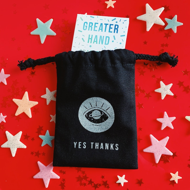 以下が含まれることがあります： 白い目のデザインと「YES THANKS」という文字が前面に印刷された黒い巾着袋。袋は赤い表面に置かれ、周りに青と白の星が散らばっています。背景には「GREATER HAND！」という文字が書かれたカードが見えます。