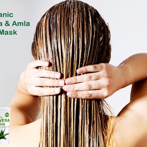 May include: A woman with wet hair applies a hair mask. Two packages of hair mask powder are visible in the foreground. The packages are labeled "Organic Amla Fruit Powder" and "Organic Aloe Vera Powder".
