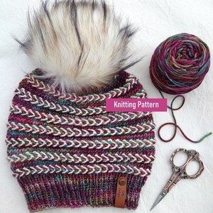 May include: A knitted hat with a white pom pom and a multi-colored yarn. The hat has a chevron pattern and a leather tag that says "Little Larkspur". The yarn is in a ball next to the hat, and there are scissors on the white surface.