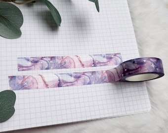 May include: Purple and pink washi tape with a marble and glitter design. The tape is rolled up and partially unrolled on a white gridded notebook.