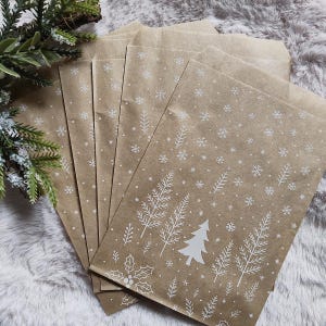 Può includere: Un gruppo di sacchetti regalo di carta marrone con disegni bianchi di fiocchi di neve e alberi. I sacchetti sono disposti a ventaglio, con un rametto di verde nell'angolo in alto a sinistra. Su una superficie morbida e strutturata.