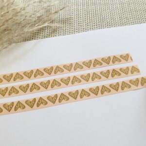 Könnte beinhalten: Eine Rolle Washi-Tape mit rosa Hintergrund und goldenen Herzen. Das Band ist abgewickelt und es liegen drei Streifen Band auf einer weißen Oberfläche.