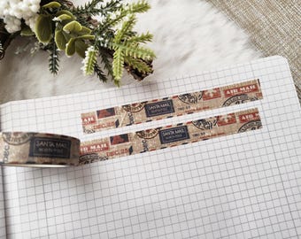 May include: Decorative washi tape with a vintage Santa Mail design. The tape features a beige background with red, black, and white accents, and the text "SANTA MAIL NORTH POLE DEC. 25". The tape is placed on a grid paper notebook.