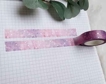 May include: A roll of washi tape with a pink and purple watercolor galaxy design and silver glitter stars. The tape is on a white gridded notebook.