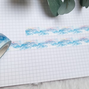 Può includere: Un rotolo di nastro washi blu e rosa con un design di montagna ad acquerello. Il nastro è parzialmente srotolato su un quaderno bianco a quadretti.