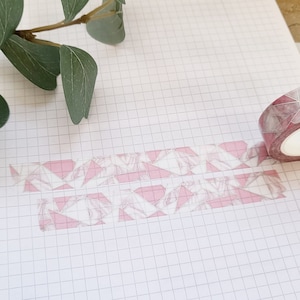 Pode incluir: Fita washi rosa e branca com um padrão geométrico. A fita está enrolada e parcialmente desenrolada em um caderno quadriculado.