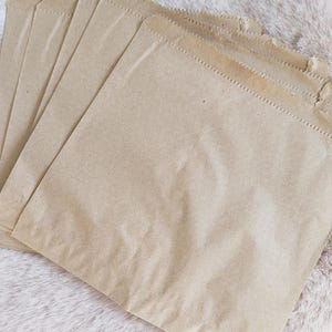 May include: A stack of five kraft paper bags, each with a rectangular shape and a folded bottom. The bags are a light brown color and have a textured surface. The top edges are sealed with a zig-zag stitch.