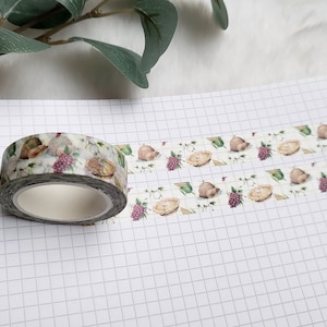 May include: A roll of decorative washi tape with a white background and a pattern of brown pies, green leaves, and purple grapes. The tape is partially unrolled on a white gridded notebook.