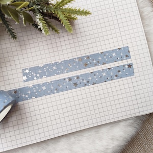 Op de afbeelding: Een rol lichtblauwe decoratieve tape met een sterrenpatroon, gedeeltelijk afgerold op een ruitjesnotitieboek. Twee stroken van dezelfde tape zijn op het papier geplakt. De tape heeft zilveren en gouden sterren.