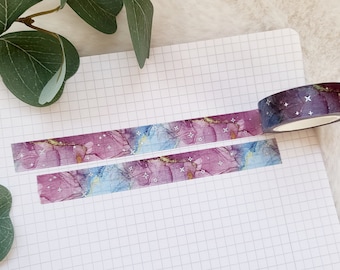May include: A roll of washi tape with a purple, blue, and gold marble pattern and silver glitter stars. The tape is unrolled on a white gridded notebook.
