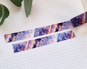 May include: Two rolls of washi tape with a galaxy print. The tape is purple, gold, and silver with a glittery finish.