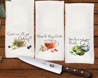 May include: Three white waffle-weave kitchen towels with tea-themed illustrations and text. One towel reads "Cant we all get Oolong?" with a tea cup. Another says "Steep & Stay Awhile" with a tea cup. The third says "You have met your Matcha". A large knife is in the foreground.